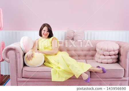 Young woman reclining on pink sofa in pastel living room wearing yellow dress and purple sandals, eyes closed in calm fashion pose 143956690