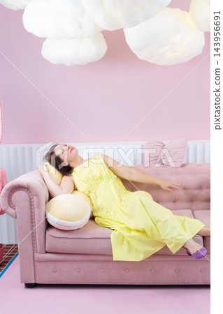 Young woman reclining on pink sofa in pastel living room wearing yellow dress and purple sandals, eyes closed in calm fashion pose Young woman reclining on pink sofa in pastel living room wearing yellow dress and purple sandals, eyes closed in calm fashion pose 143956691