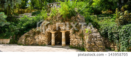 Man-made stone grotto with classical columns in a lush green park in Pyatigorsk.. Artificial cave entrance with pillars and overgrown foliage. Historical garden landscape architecture in summer Man-made stone grotto with classical columns in a lush green park in Pyatigorsk.. Artificial cave entrance with pillars and overgrown foliage. Historical garden landscape architecture in summer 143958705