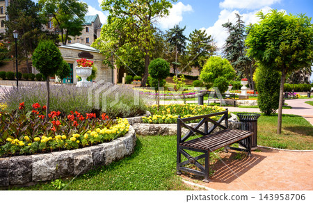 Landscaped public park with a wooden bench and colorful flower beds. Urban garden featuring topiary trees, stone paths, and green lawns on a sunny summer day in Pyatigorsk, Russia Landscaped public park with a wooden bench and colorful flower beds. Urban garden featuring topiary trees, stone paths, and green lawns on a sunny summer day in Pyatigorsk, Russia 143958706