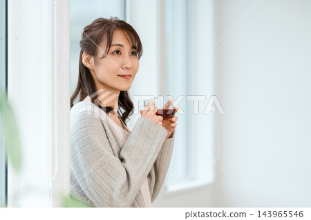 A woman in loungewear relaxing by the window with a cup of tea. 143965546