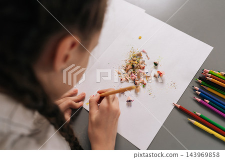 View from shoulder of schoolgirl drawing with colored pencil on white paper, surrounded by colorful sharpened pencils, wood shavings, promoting childhood creativity and artistic learning at school. View from shoulder of schoolgirl drawing with colored pencil on white paper, surrounded by colorful sharpened pencils, wood shavings, promoting childhood creativity and artistic learning at school. 143969858
