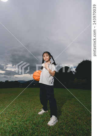 Young girl standing on green field holding orange basketball with cloudy sky and trees in background confidently posing 143970039