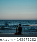 male photographer kneeling on sandy beach capturing ocean waves at dusk, man holding camera near sea water during dark twilight 143971282
