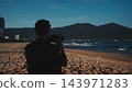 male photographer shooting pictures on sandy beach facing dark mountains, silhouette of man capturing coastal landscape near ocean waves during dusk 143971283