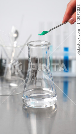 Scientist adding green powder into Erlenmeyer flask, conducting chemical experiment in research laboratory Scientist adding green powder into Erlenmeyer flask, conducting chemical experiment in research laboratory 143973828