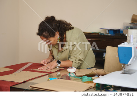 Focused woman pins a sewing pattern onto red fabric while creating handmade fashion designs at a home tailoring workshop table 143974851