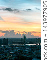 As day transitions to night, a warm golden glow illuminates the sky above the Chao Phraya River, casting dramatic silhouettes of distant storm clouds over the illuminated Kasikornbank Head Office 143977905
