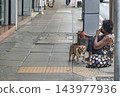 An elderly woman wearing a face mask sits on a Bangkok sidewalk begging for money with her small dog on a leash. 143977936