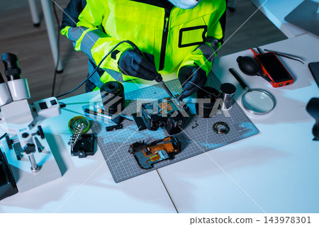 Forensic technician in a high visibility jacket examines a dismantled camera with tools, microscope, evidence items and a tablet on a laboratory desk. Forensic technician in a high visibility jacket examines a dismantled camera with tools, microscope, evidence items and a tablet on a laboratory desk. 143978301