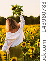 Woman enjoying a quiet moment in a blooming sunflower field while lifting a flower toward the warm evening sunlight. The serene outdoor scene reflects tranquility, mindfulness and rural summer 143980783