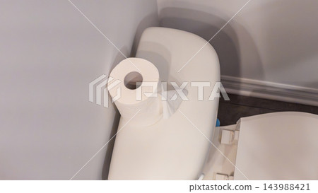 White toilet paper roll resting on a toilet tank beside a partially visible open lid in a simple indoor bathroom setting with wall trim and ceramic fixture White toilet paper roll resting on a toilet tank beside a partially visible open lid in a simple indoor bathroom setting with wall trim and ceramic fixture 143988421