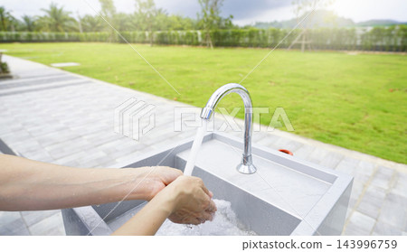 Woman washing hands with clean running tap water at an outdoor sink beside a green lawn on a sunny day, promoting personal hygiene, sanitation, health protection and responsible water use. Woman washing hands with clean running tap water at an outdoor sink beside a green lawn on a sunny day, promoting personal hygiene, sanitation, health protection and responsible water use. 143996759
