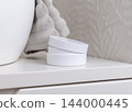 Open white jar mockup with a lid near grey towel in beige bathroom. Unbranded cosmetic packaging 144000445