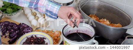 Hands add dried hibiscus, also called Sudanese rose, to a pot of dye for Easter eggs in a home kitchen, with turmeric used as another natural dye, banner 144001997