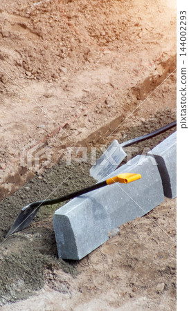 Installation process of road curbing, with grey concrete curb stones, shovel, and trowel visible in dug trench on construction site, preparation work for infrastructure project, vertical photo 144002293
