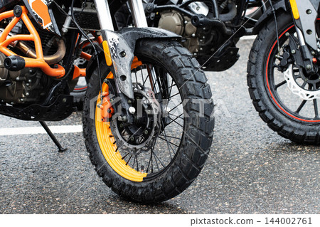 Close up view front wheel orange black adventure motorcycle parked wet asphalt street, textured off-road tire visible brake disc caliper components, bright day light Close up view front wheel orange black adventure motorcycle parked wet asphalt street, textured off-road tire visible brake disc caliper components, bright day light 144002761