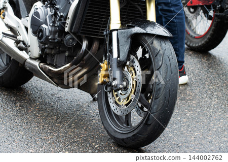 Close up view of front wheel motorcycle, tire tread on wet asphalt, golden brake caliper, front disc brake system, engine detail, exhaust pipes, shiny suspension forks, showcasing modern vehicle Close up view of front wheel motorcycle, tire tread on wet asphalt, golden brake caliper, front disc brake system, engine detail, exhaust pipes, shiny suspension forks, showcasing modern vehicle 144002762