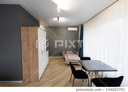Modern studio apartment interior showing compact living room space with contemporary dining table, black chairs, wardrobe cabinet, sofa bed, parquet floor, tulle curtains Modern studio apartment interior showing compact living room space with contemporary dining table, black chairs, wardrobe cabinet, sofa bed, parquet floor, tulle curtains 144002791
