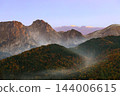 Autumn forested hills and rugged alpine peaks emerge through mist in golden morning light 144006615