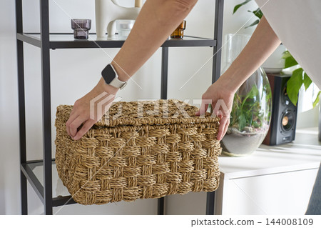 Close up of woman placing large woven wicker storage basket with lid on shelf Close up of woman placing large woven wicker storage basket with lid on shelf 144008109