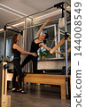 A Pilates instructor supports a client through a side stretch on a trapeze table in a modern studio 144008549