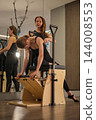 A Pilates coach assists a client with a deep forward movement on studio equipment in front of mirrors 144008553