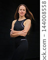 A woman in black sportswear poses with her arms crossed against a dark wall 144008558