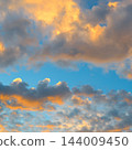 Scenic cloudscape of fluffy orange clouds glowing under warm evening sun light against blue sky in square view 144009450