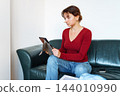 young woman using tablet while sitting on leather green sofa 144010990