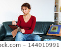 young woman using tablet while sitting on leather green sofa 144010991