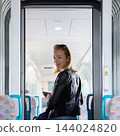 Young blond woman in jeans, shirt and leather jacket holding her smart phone and purse while riding modern speed train arriving to final train station stop. Travel and transportation 144024820