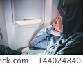 Tired blonde casual caucasian woman wearing sporty hoodie napping on seat while traveling by airplane. Commercial transportation by planes. Authentic image of real people 144024840