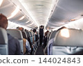 Interior of airplane with passengers on seats and stewardess in uniform walking the aisle, serving people. Commercial economy flight service concept 144024843