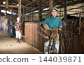 Portrait of an woman in stable with bridle and saddle in her hands 144039871