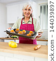 Smiling senior woman in apron holding sheet pan cupcakes 144039897