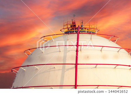Spherical gas storage tank with red piping beneath an orange cloudy sky at a petrochemical facility, illustrating industrial fuel storage and energy infrastructure with a dramatic atmospheric backdrop Spherical gas storage tank with red piping beneath an orange cloudy sky at a petrochemical facility, illustrating industrial fuel storage and energy infrastructure with a dramatic atmospheric backdrop 144041652