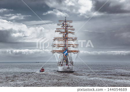 Majestic three masted tall ship sailing forward on deep blue ocean under dramatic dark storm clouds sky horizon Majestic three masted tall ship sailing forward on deep blue ocean under dramatic dark storm clouds sky horizon 144054689